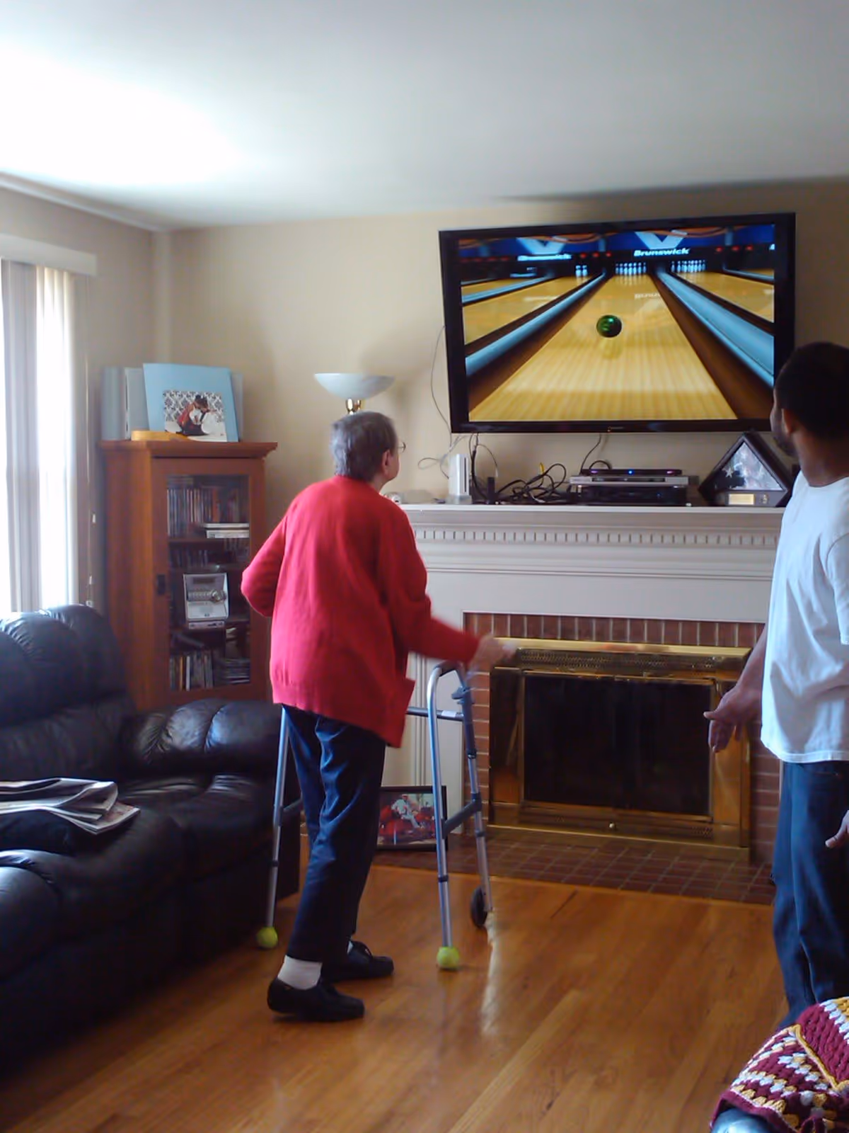 An elderly person using a walker plays a bowling video game on a wall-mounted TV in a living room while another person watches.