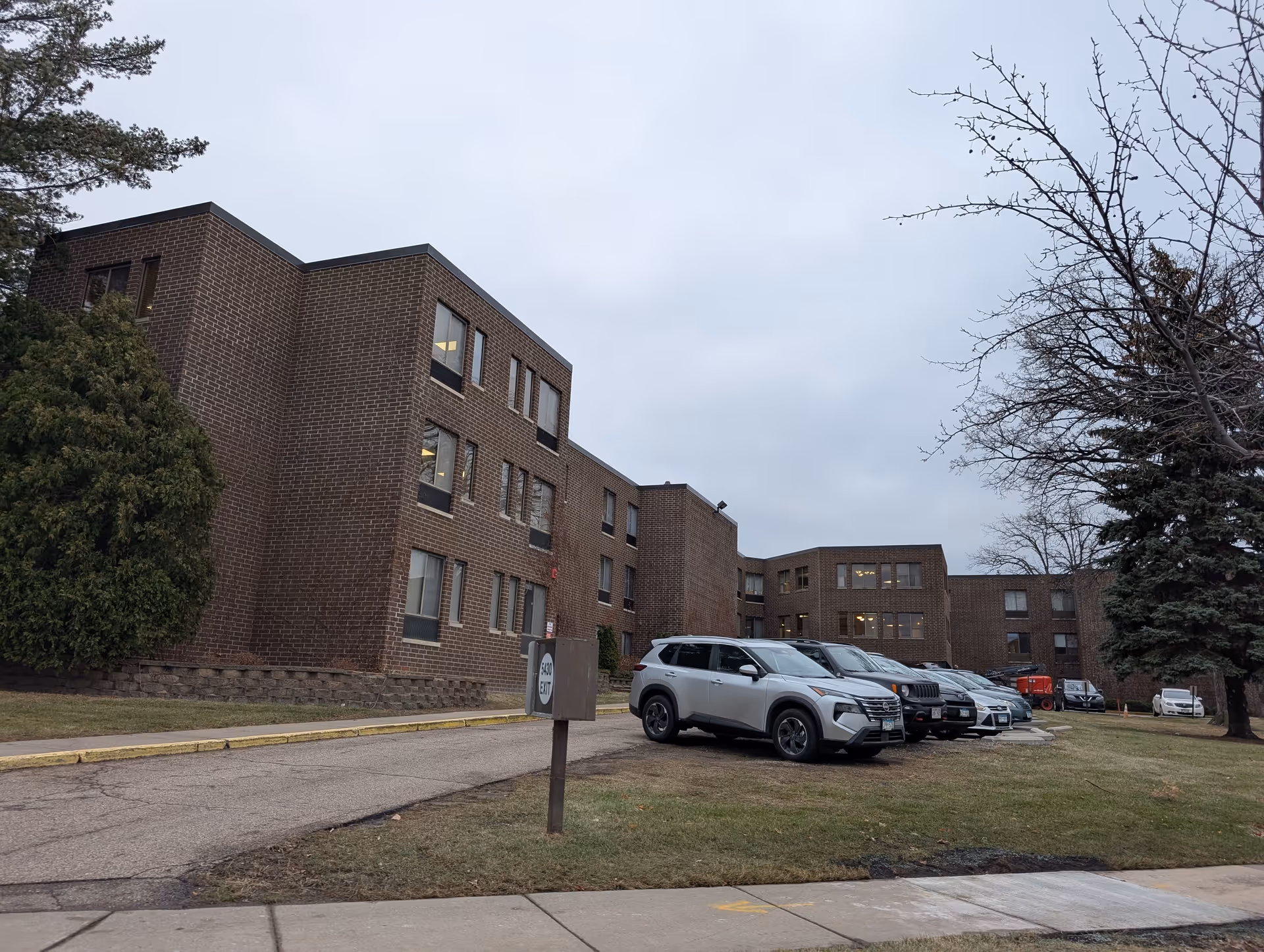 Brick three-story senior living facility building with a row of parked cars on the lawn under an overcast sky.