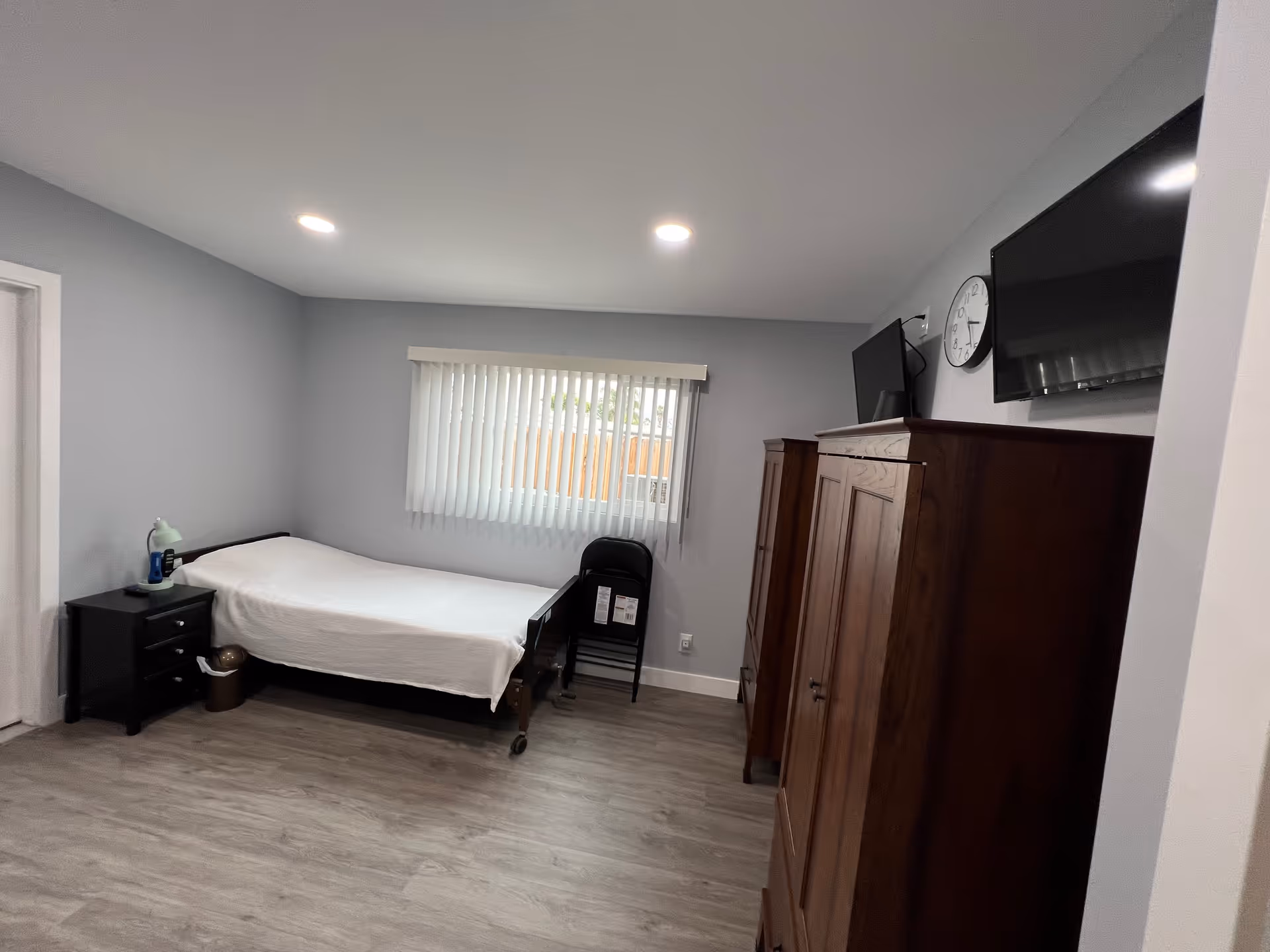 Small single-occupancy bedroom with a bed and nightstand on the left, two wooden wardrobes and a wall-mounted TV on the right, and a window with vertical blinds.
