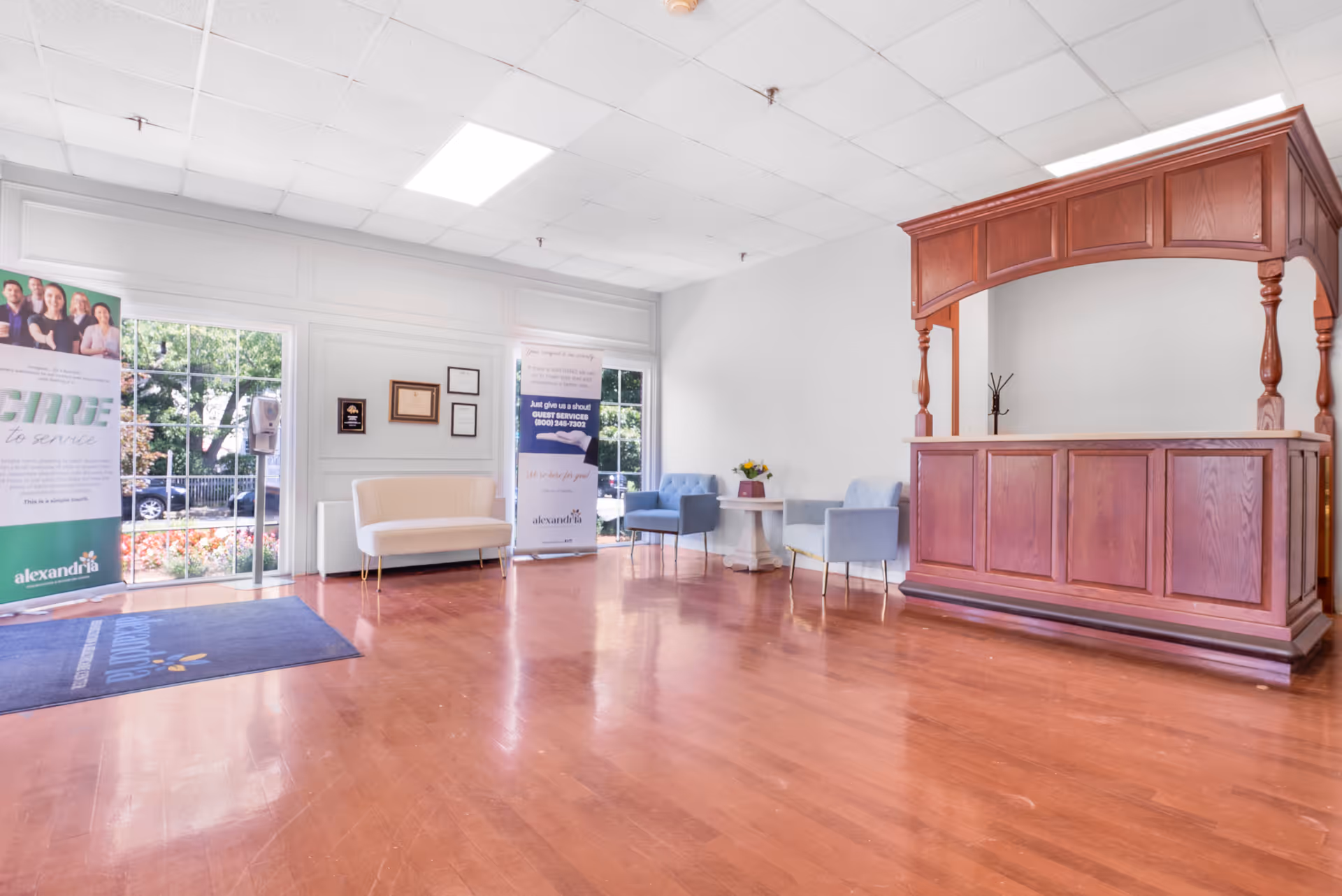 A bright and spacious reception area with wooden flooring, a large wooden reception desk on the right, a white couch and two blue chairs around a small table with flowers, large windows letting in natural light, and promotional banners near the entrance.