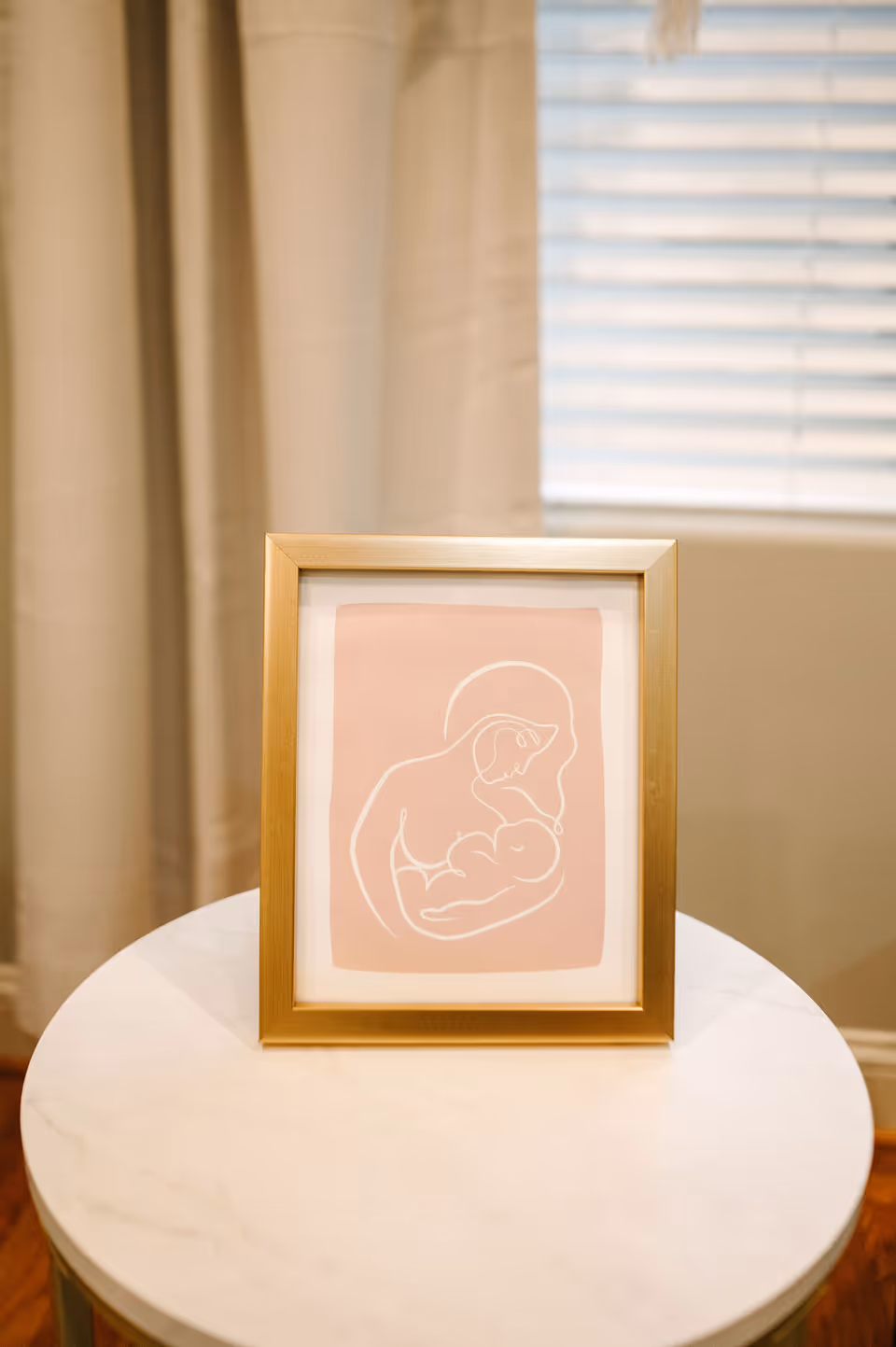 Framed pink line-art of a mother breastfeeding on a round marble-top table indoors.