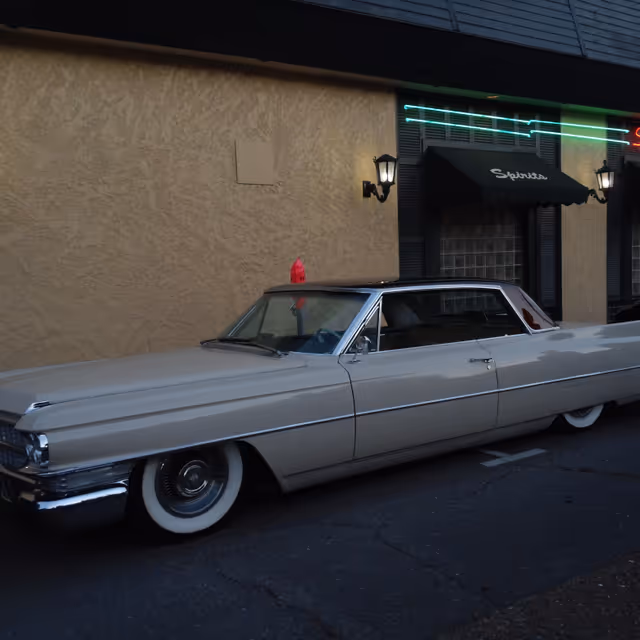 A vintage white car parked beside a beige building with black awnings and neon signage.