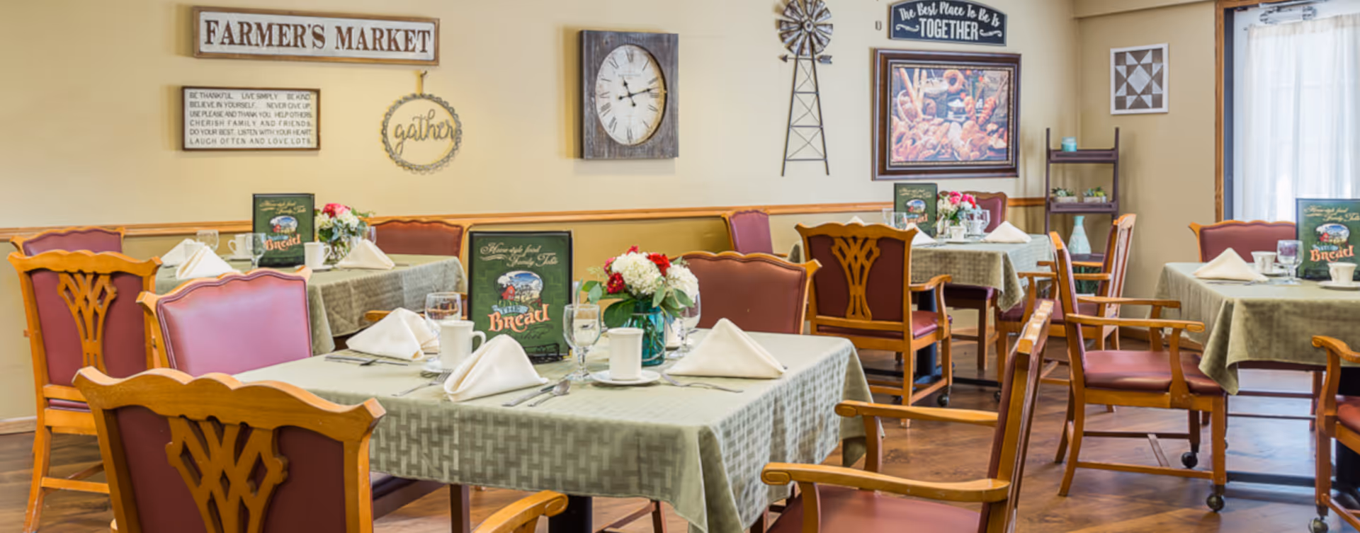 A cozy dining room with several tables covered in light green tablecloths, each set with white napkins, cups, glasses, and silverware. The chairs have wooden frames with reddish upholstery. The walls are decorated with various signs, including one that says 'FARMER'S MARKET,' a clock, a windmill decoration, and framed artwork. There are also flower arrangements on the tables and a window with sheer white curtains letting in natural light.