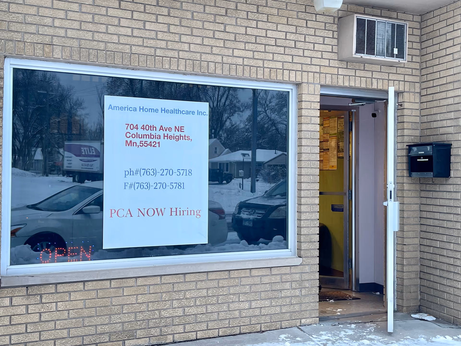 Exterior view of a brick building with a large window, open entrance, and hiring sign.