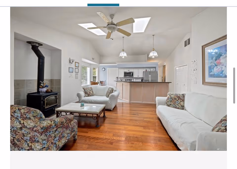 A bright living room with wooden floors, a white sofa, a floral armchair, a white loveseat, a coffee table, a ceiling fan, and a wood-burning stove. The room opens up to a kitchen area with a breakfast bar, pendant lights, and stainless steel appliances.