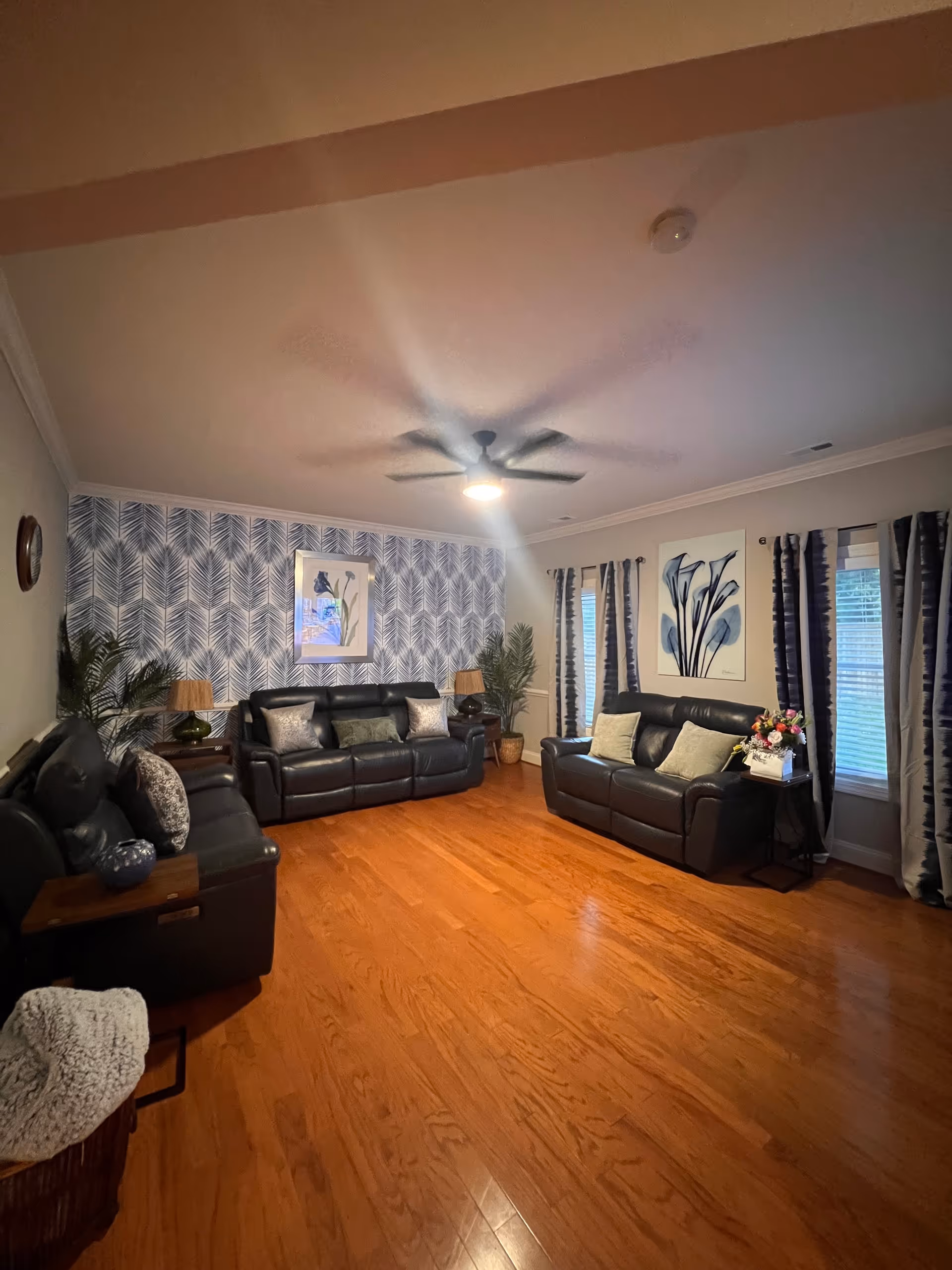 A spacious living room with leather sofas, wooden floors, plants, and patterned wallpaper.