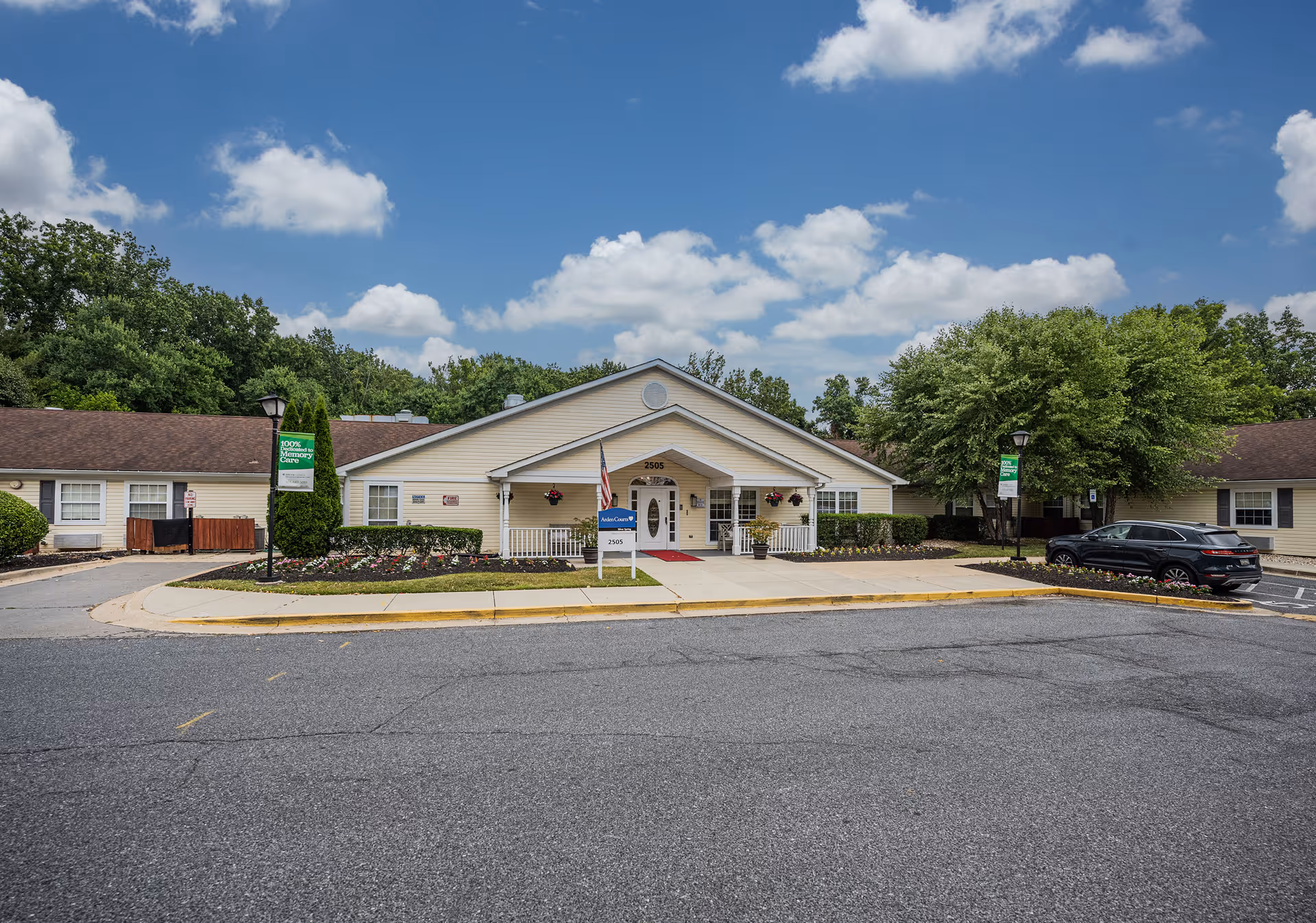 Exterior view of Arden Courts - ProMedica Memory Care Community building in Silver Spring, featuring a single-story structure with a beige facade, a peaked roof, and a central entrance with a small porch. There are flower beds and shrubs in front, a parking area with a black car on the right, and a partly cloudy blue sky overhead.