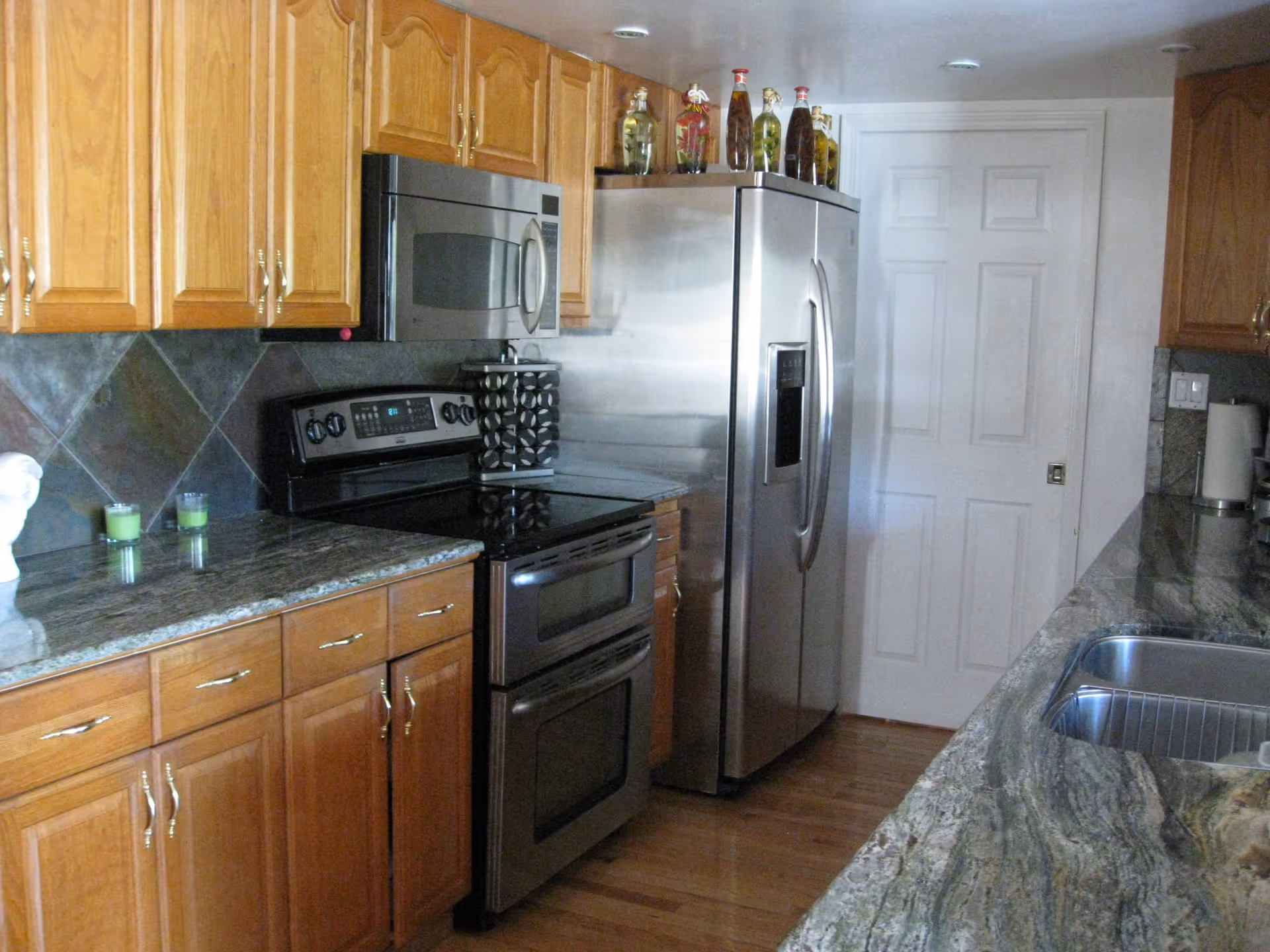 A kitchen with wooden cabinets, granite countertops, a stainless steel refrigerator, a stainless steel microwave above a black stove, and a double sink. There are decorative bottles on top of the refrigerator and two green candles on the counter.