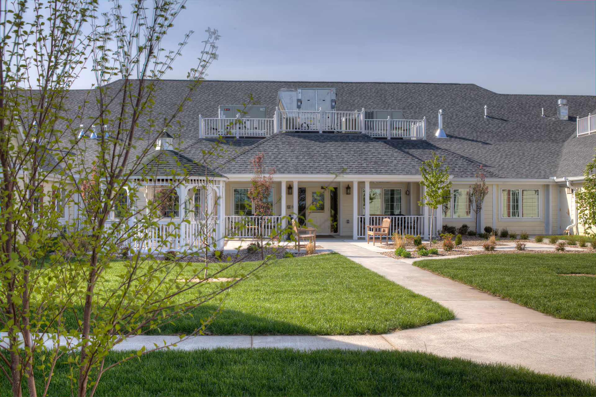 Front exterior view of Homestead Assisted Living of Albia showing a single-story building with a gray shingled roof, white railings, and a covered porch area with chairs. There is a well-maintained green lawn and a concrete walkway leading to the entrance, with young trees and shrubs planted around the building.
