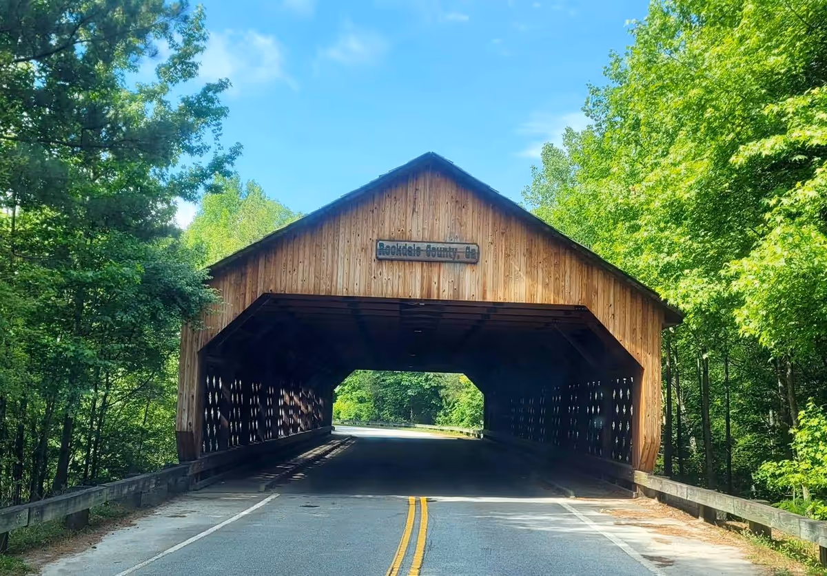 Wooden covered bridge spans a road through a forest of green trees.