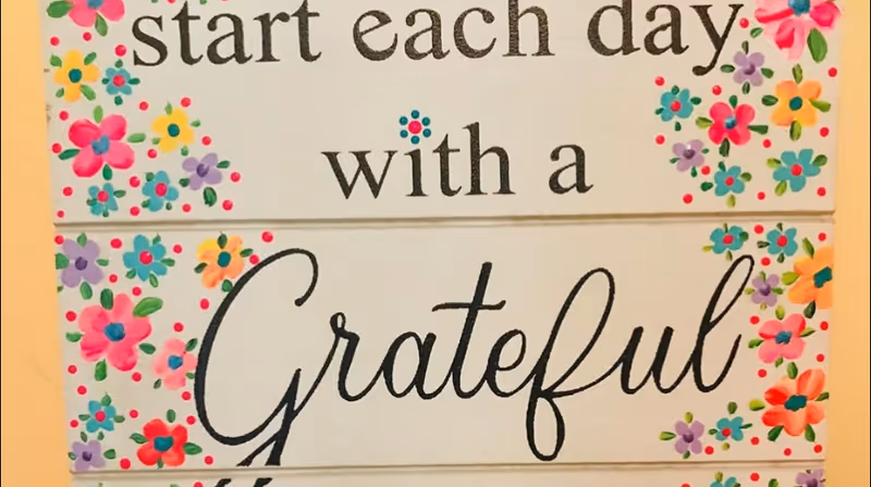 Decorative wooden sign with a floral border and the message start each day with a Grateful.