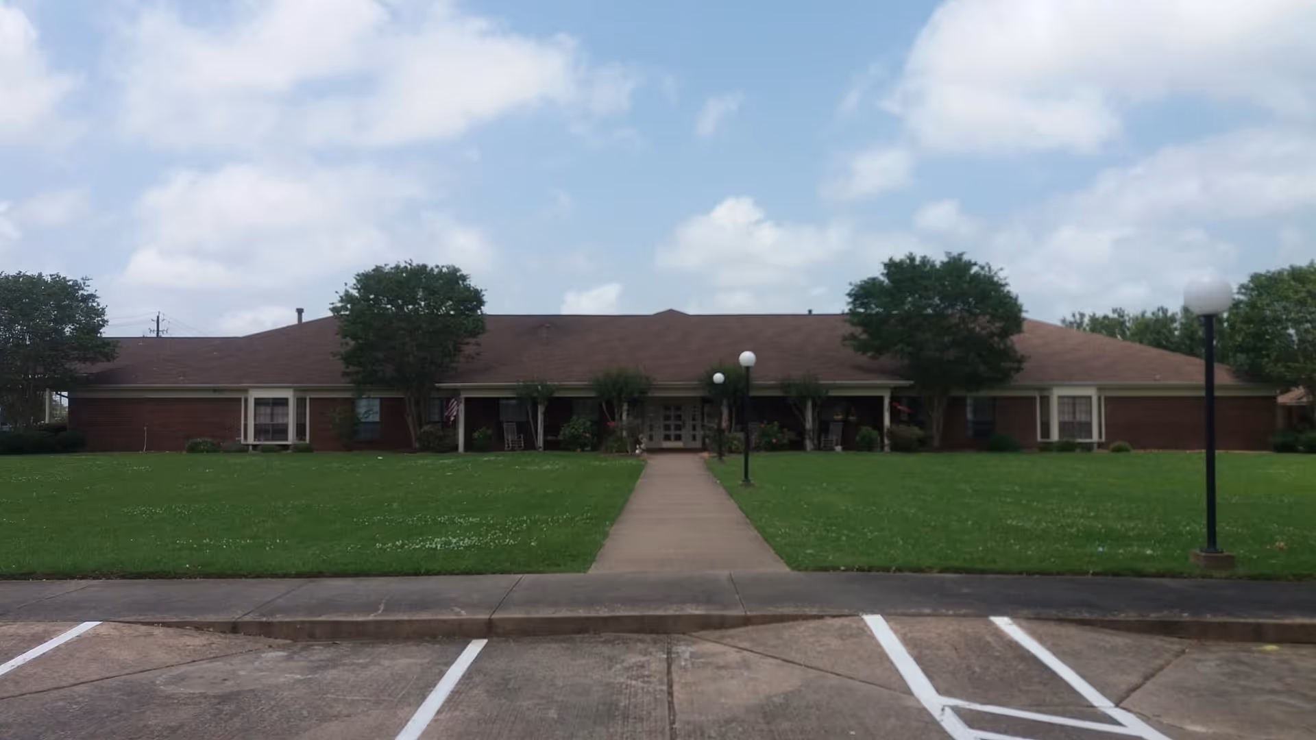 Front exterior of a single-story brick senior living facility with a wide lawn, central walkway, and lamp posts.