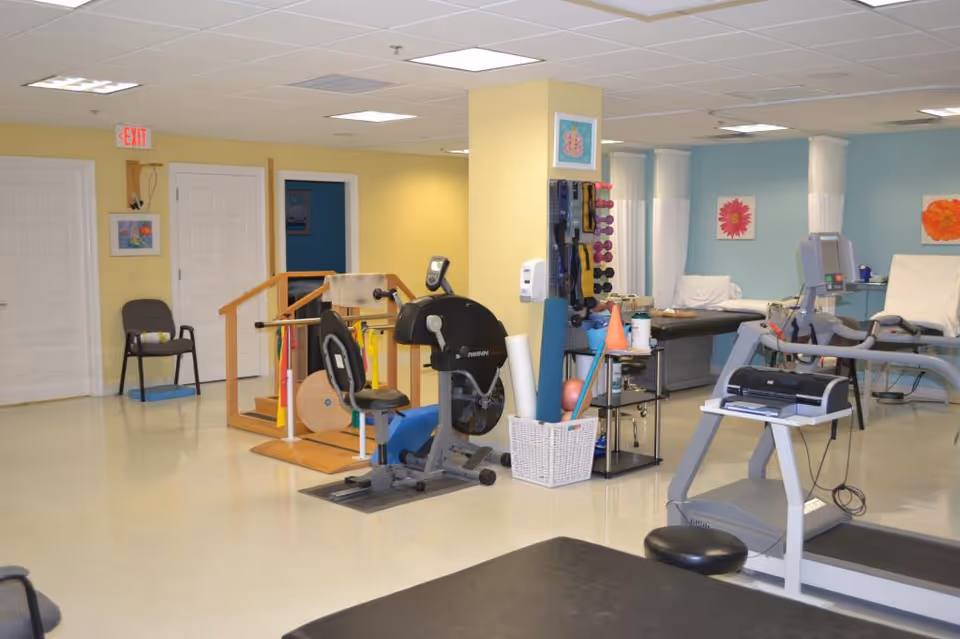 Bright rehabilitation gym with a treadmill, stationary bike, weights, and seating in a senior living facility.