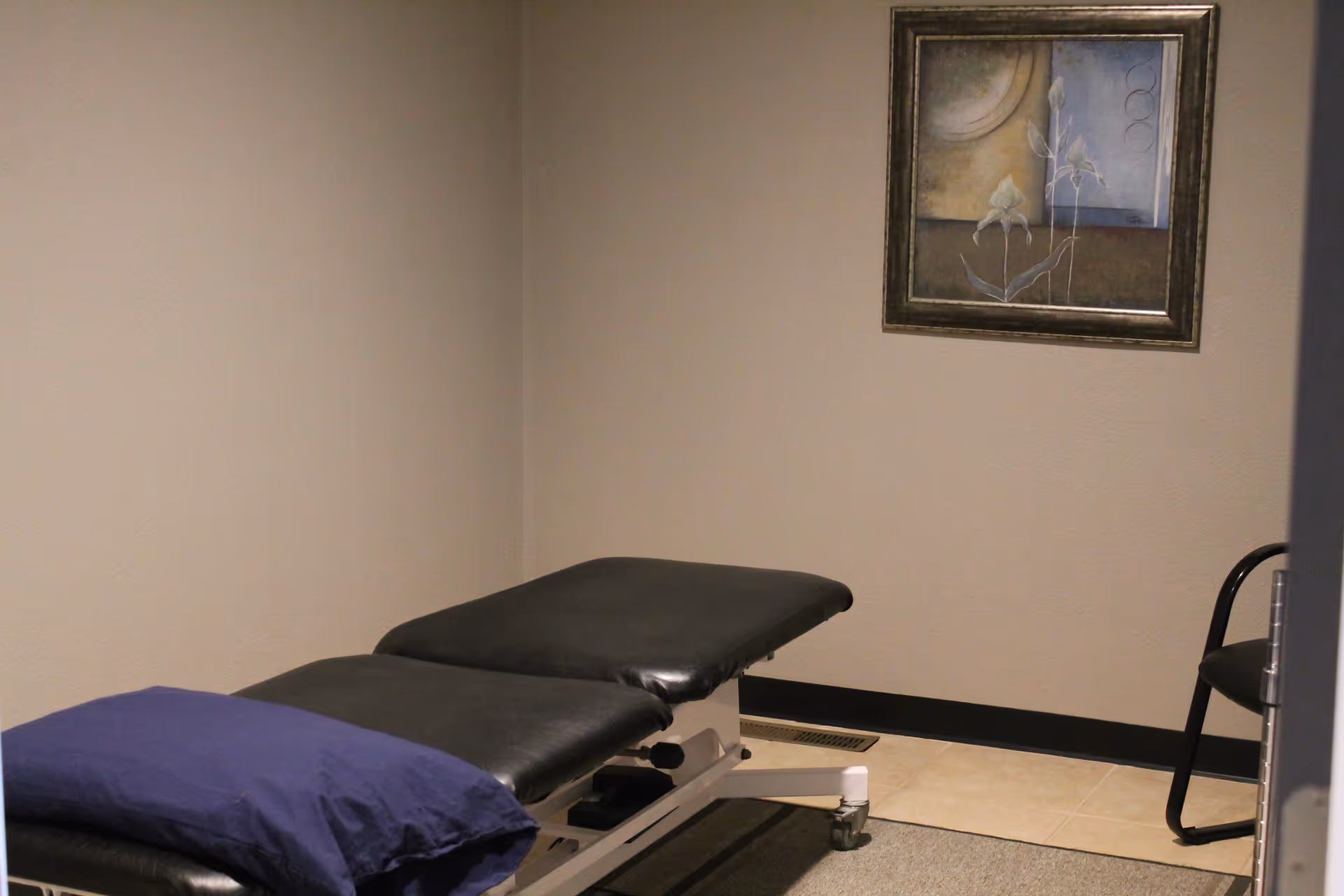An examination table in a small therapy room with a framed painting on the beige wall.