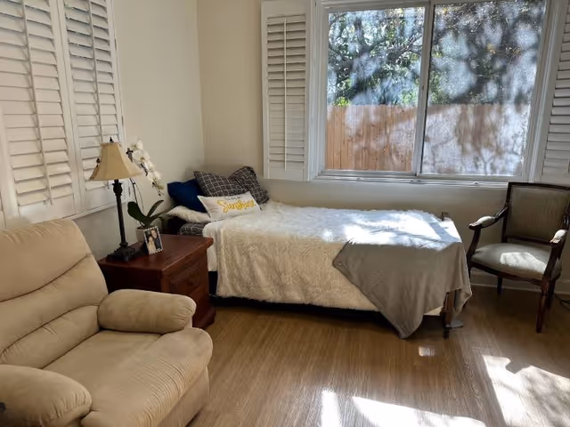 Sunny room with a single bed by a large window, a recliner, nightstand with lamp and a chair.