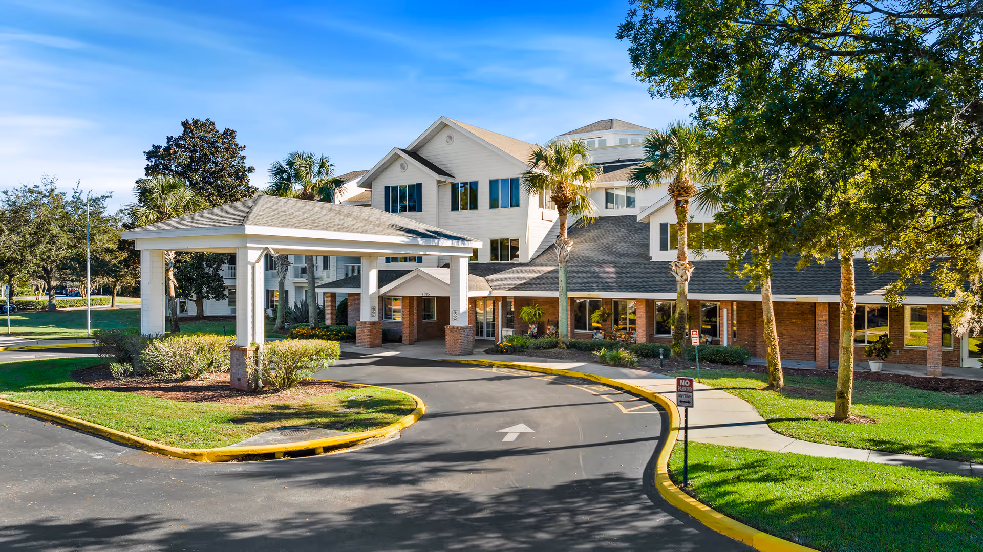 Exterior view of The Tremont - A Provincial Senior Living Community building with a covered entrance driveway, surrounded by green lawns, palm trees, and other trees under a clear blue sky.