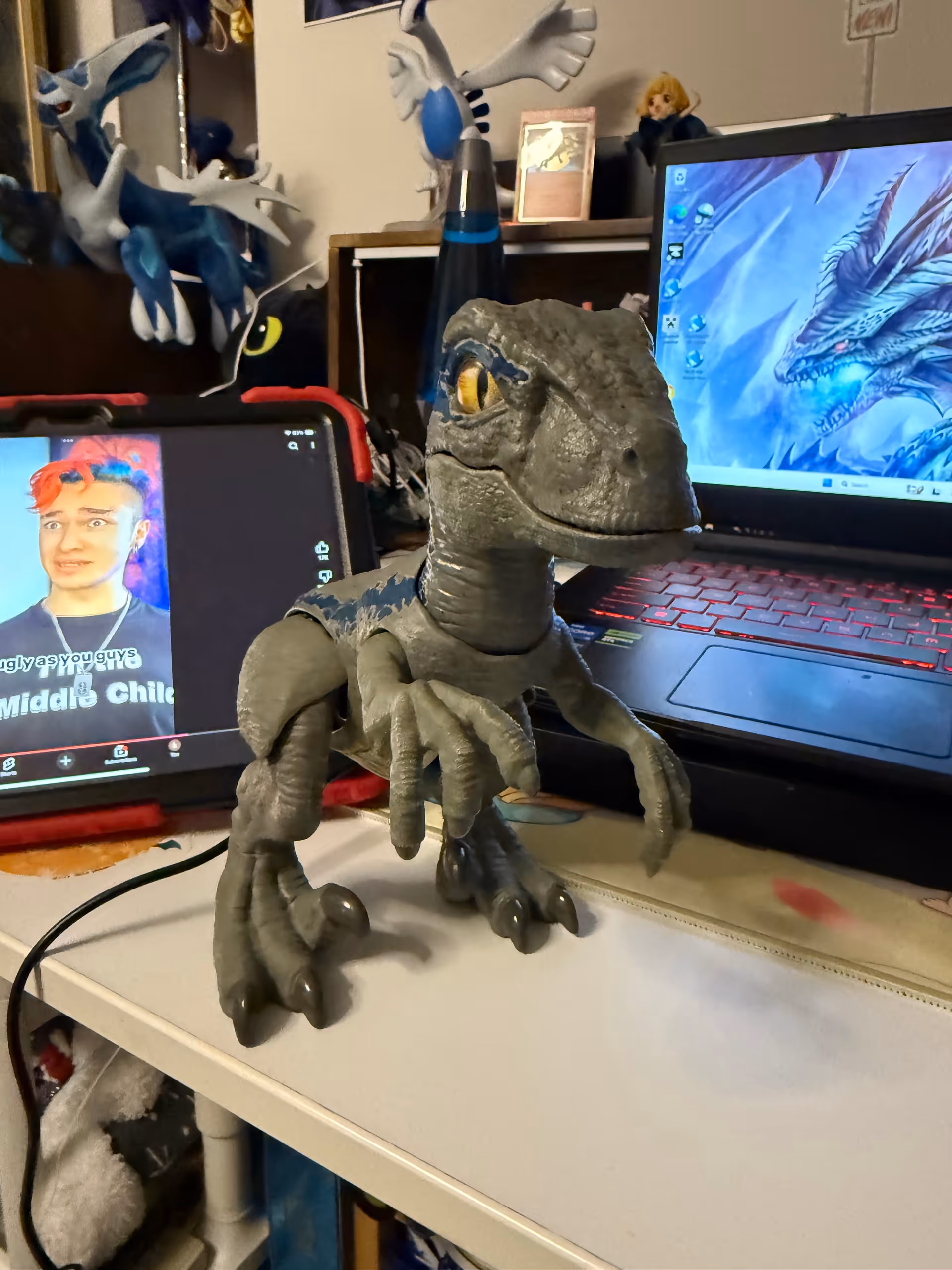 Dinosaur toy on a desk with a tablet and laptop in a cluttered interior room
