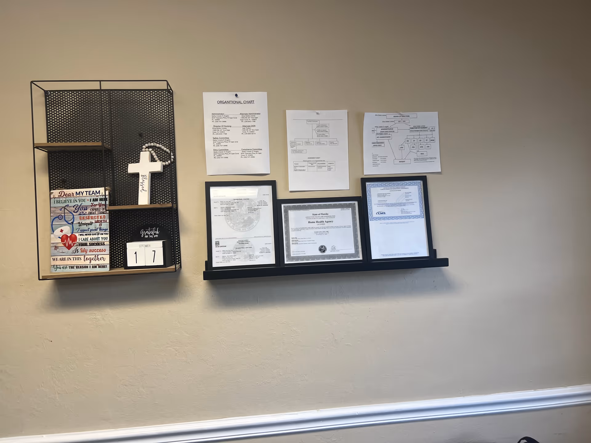 Beige wall with metal shelf displaying a cross, charts, and framed certificates.