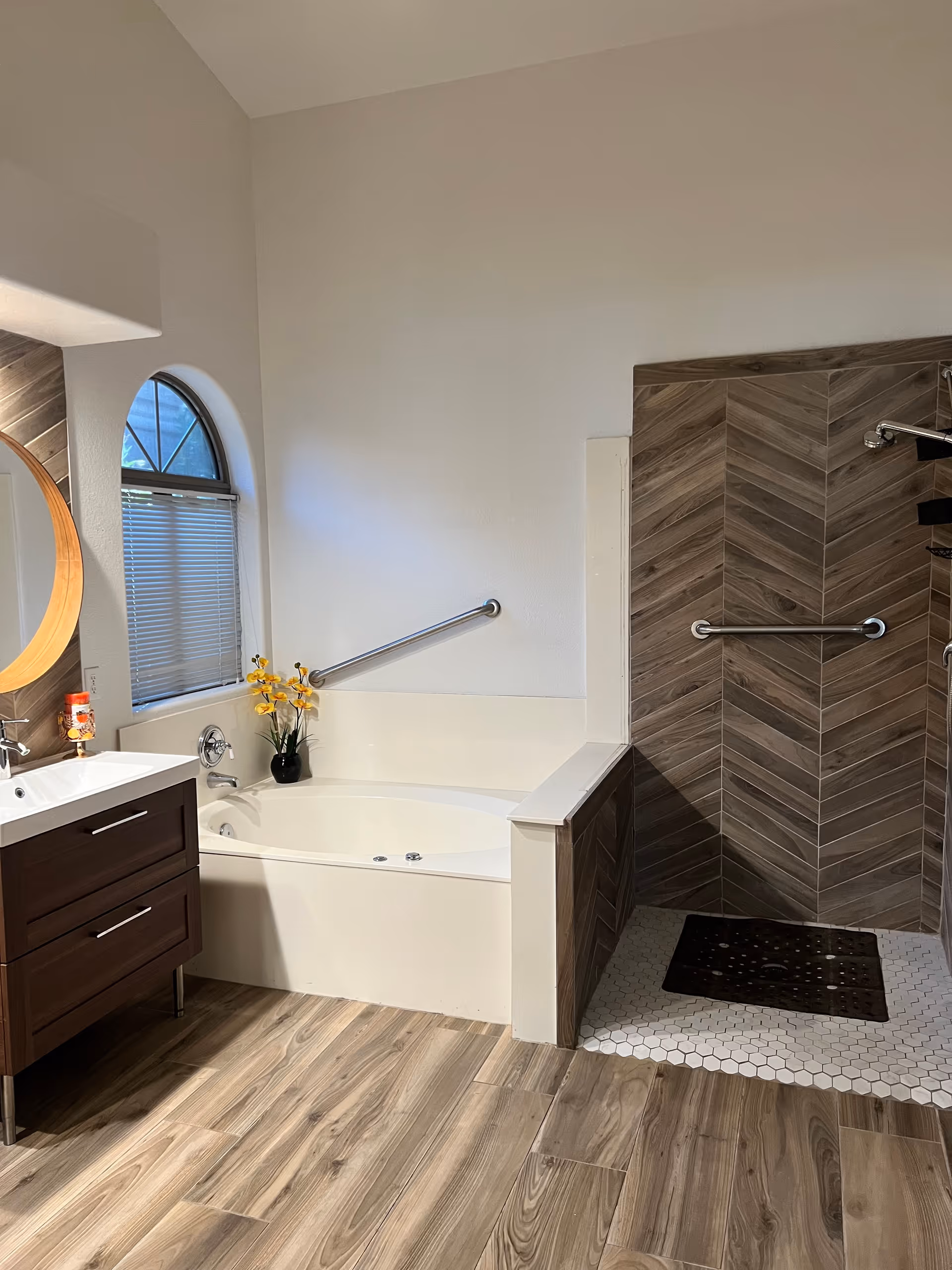 A modern bathroom featuring a white bathtub with a safety grab bar, a walk-in shower with wood-patterned tiles and a grab bar, a wooden vanity with a white sink, a round mirror, a small window with blinds, and a vase with yellow flowers on the bathtub edge. The floor has wood-like tiles.
