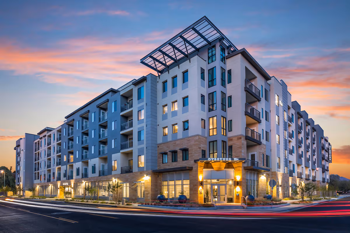 Exterior view of a modern multi-story senior living facility named Overture Kierland at dusk, with lights glowing from the windows and a colorful sunset sky in the background.