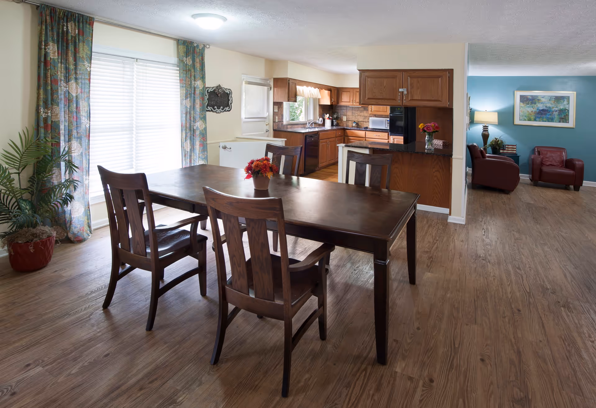 A dining area with a dark wooden table and four matching chairs, a small flower arrangement on the table, large window with floral curtains, adjacent kitchen with wooden cabinets and black countertops, and a living room with blue walls, red armchairs, a lamp, and a framed painting.