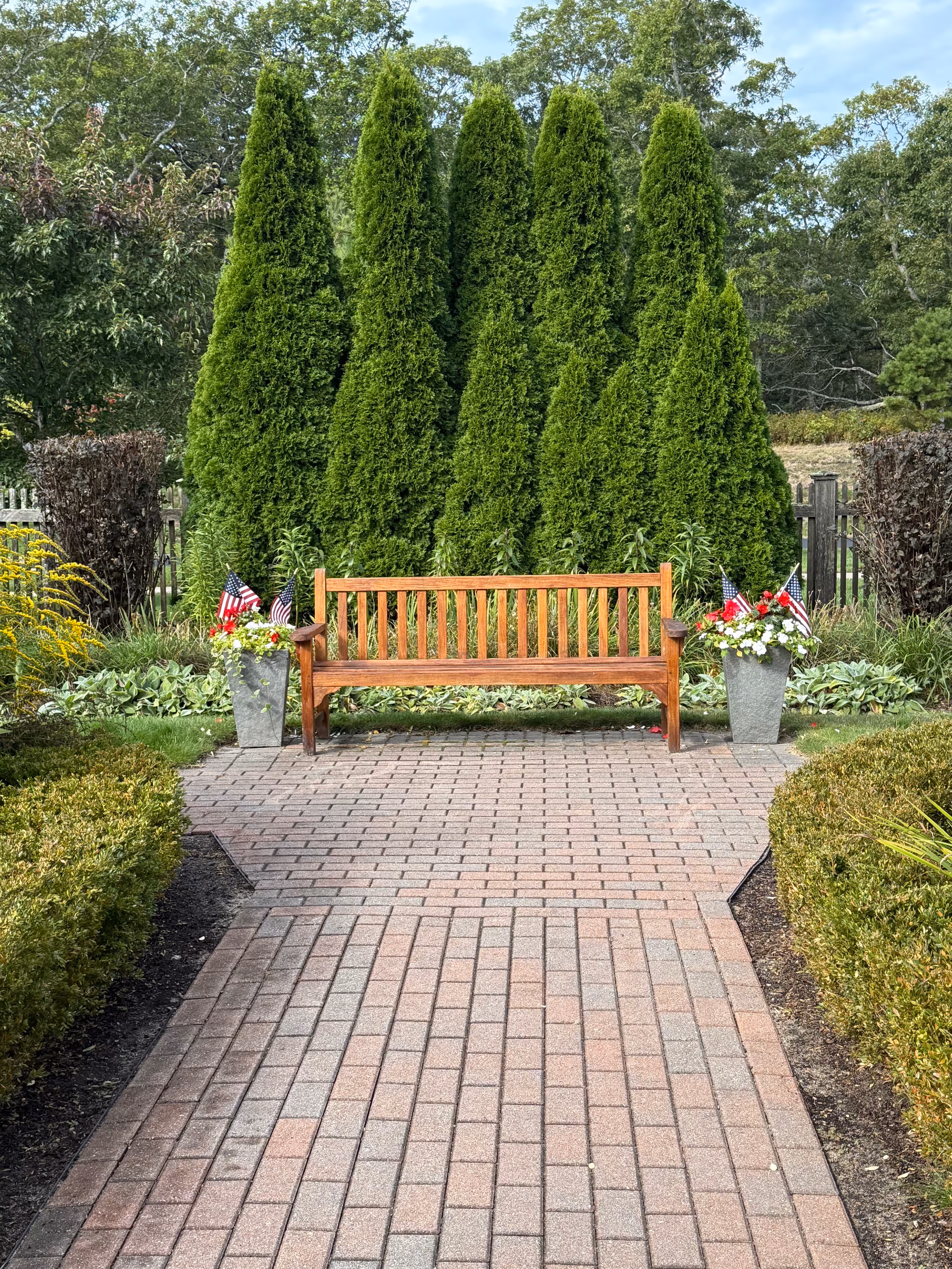 Wooden bench sits along a brick path, bordered by evergreens and patriotic planters.