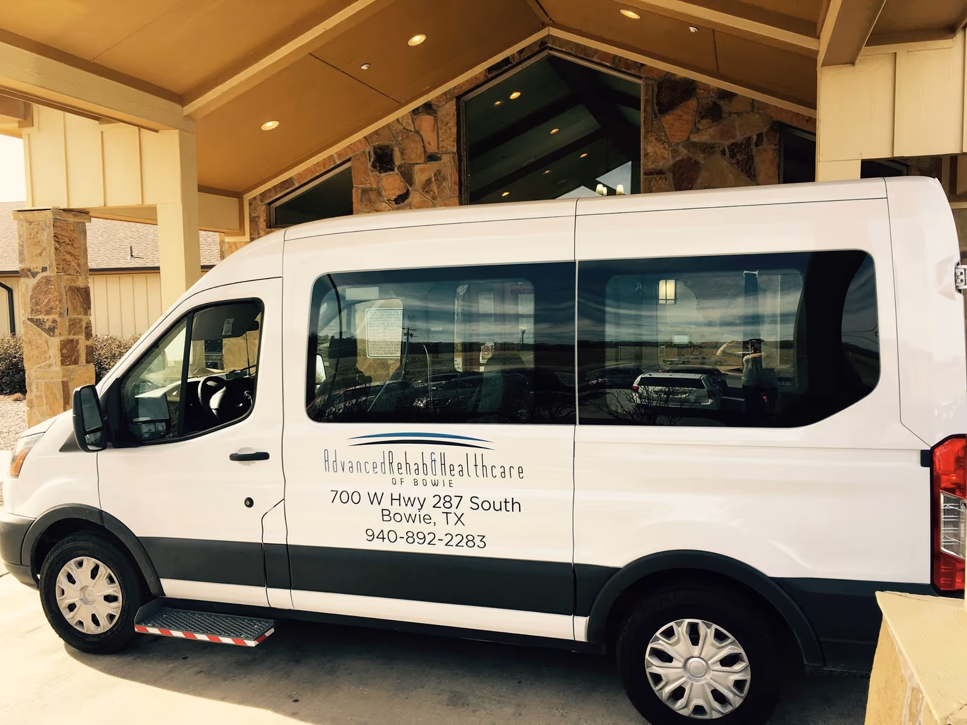A white passenger van parked under a covered entrance with stone pillars. The van has signage on the side that reads 'Advanced Rehab & Healthcare of Bowie, 700 W Hwy 287 South, Bowie, TX, 940-892-2283'.