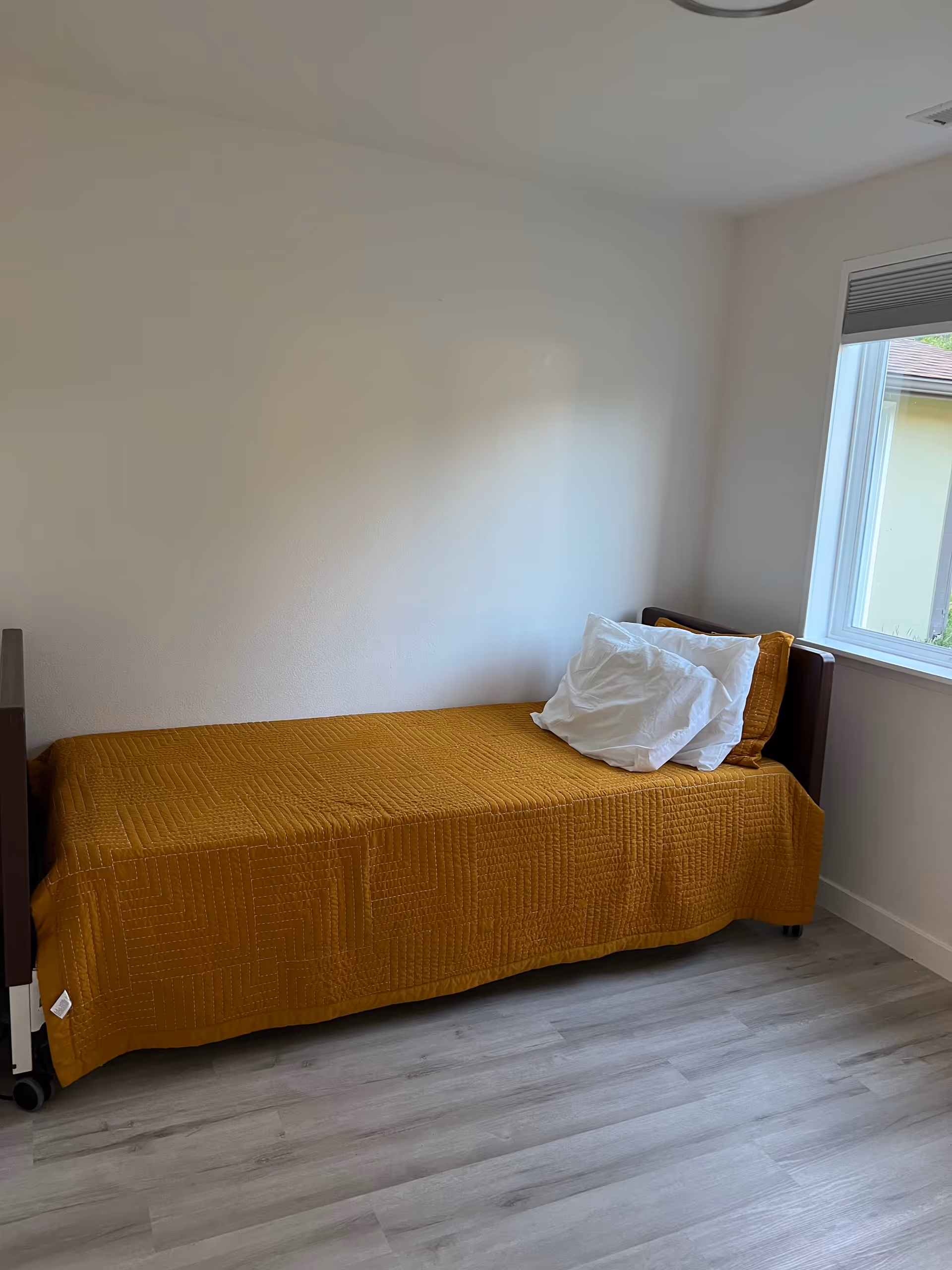 A single bed with mustard quilt beside a window in a bright, minimal bedroom.