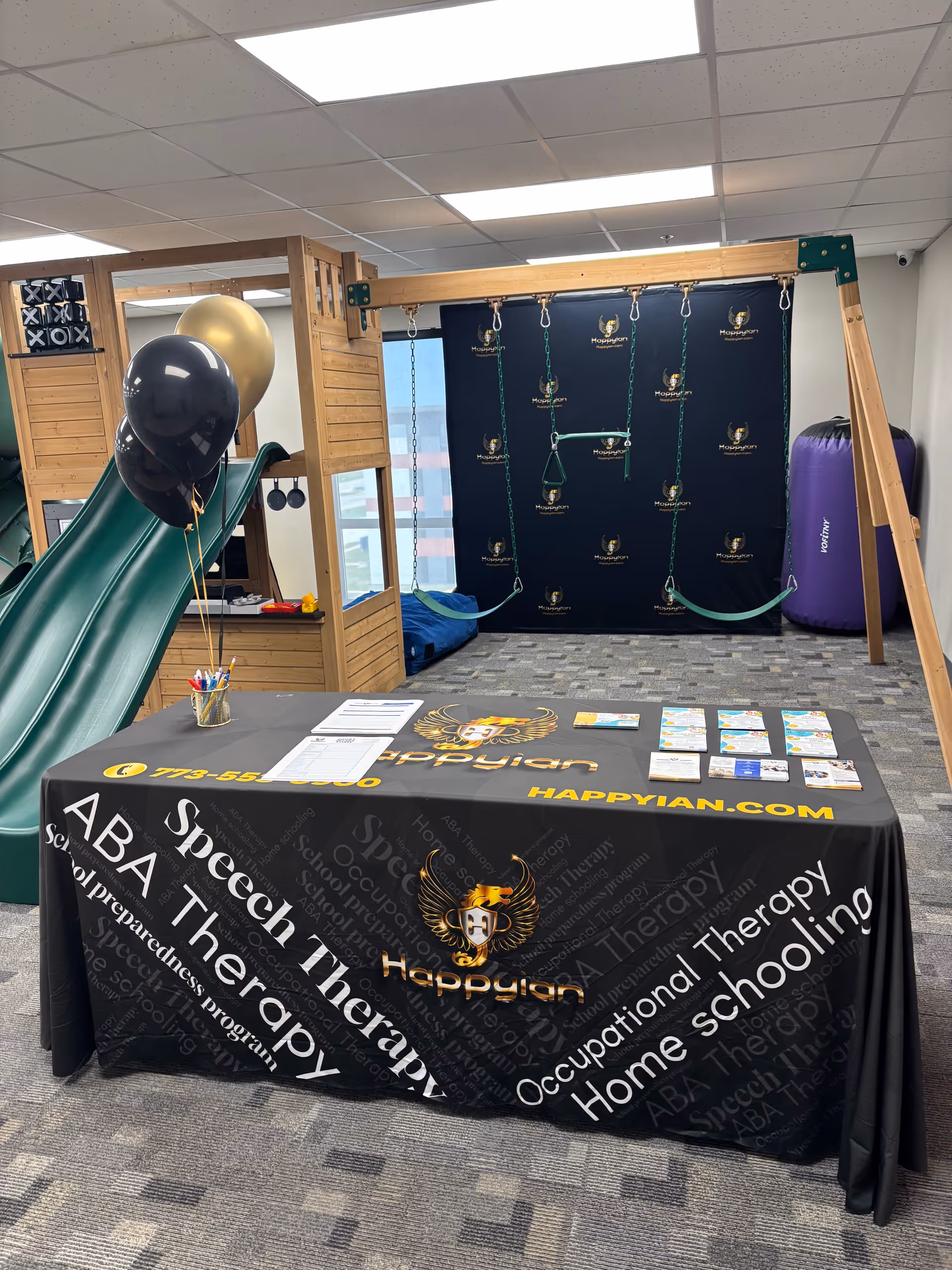 Indoor play area with a wooden climbing frame, swings, balloons, and a promotional information table.