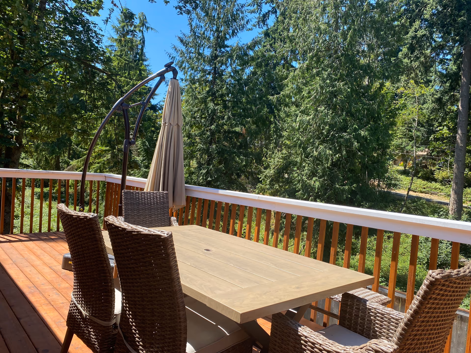 Outdoor wooden deck with a rectangular table and six wicker chairs, surrounded by a wooden railing. There is a closed beige patio umbrella attached to a curved stand. The background features tall green trees under a clear blue sky.