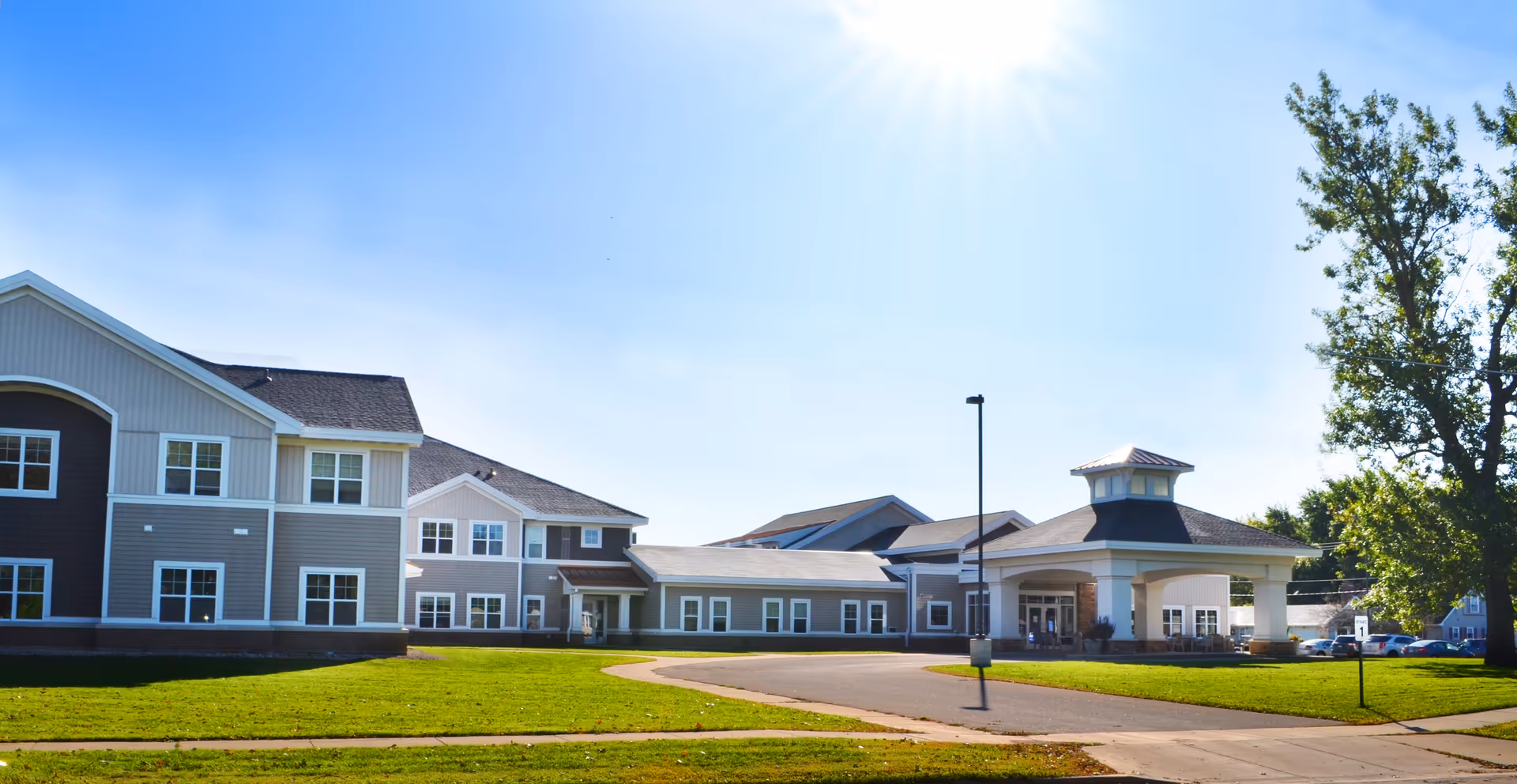 Exterior view of St. Mark's Living Assisted Living & Memory Care facility on a sunny day, showing a large building with multiple windows, a covered entrance, green lawn, and a tree on the right side.