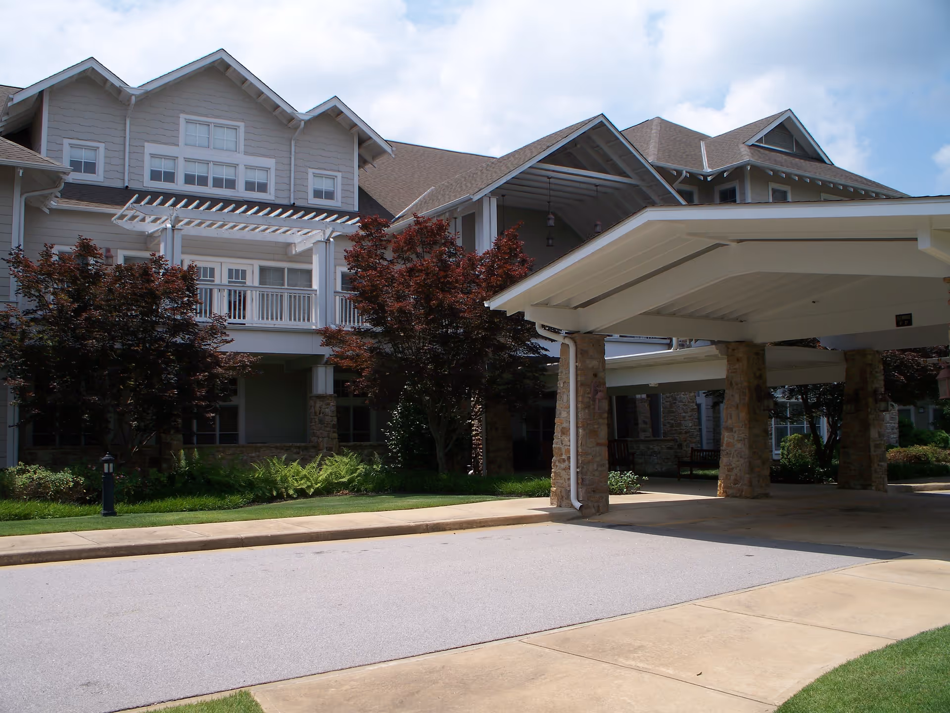 Exterior view of a senior living facility named Vernon Woods showing a covered driveway entrance with stone pillars, landscaped greenery, and a multi-story building with balconies and large windows under a partly cloudy sky.