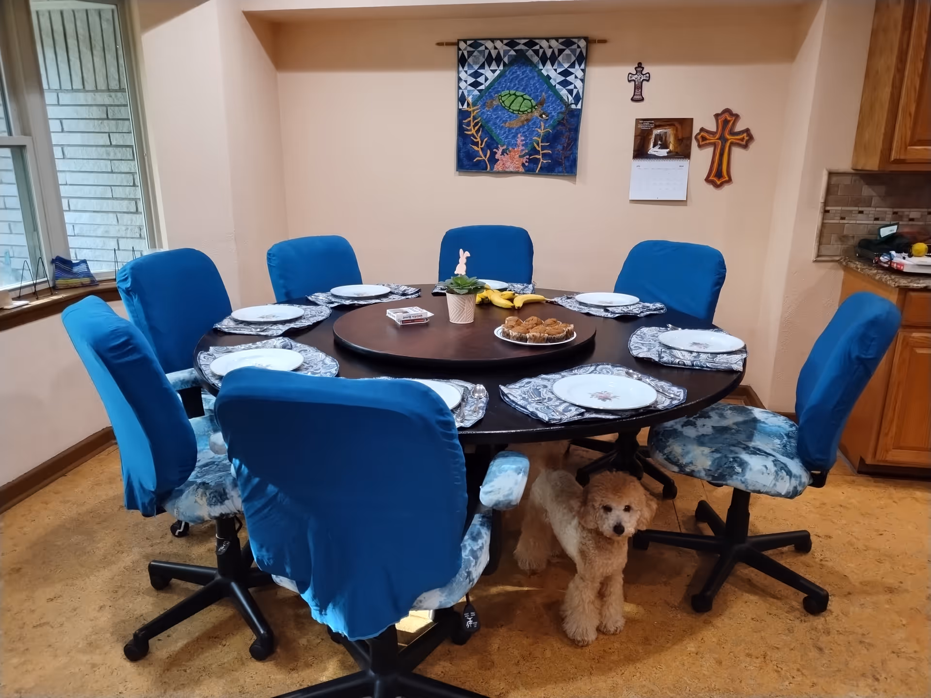Round dining table with blue chairs arranged for a meal; small dog under the table.