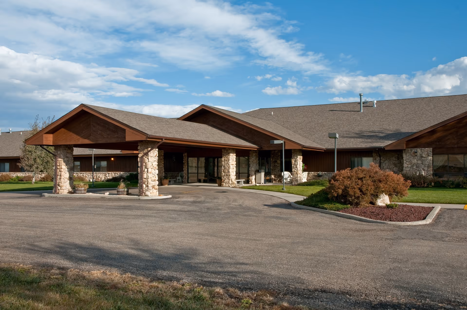 Single-story senior living facility with a covered stone-pillared entrance, driveway, and landscaped lawn under a blue sky.