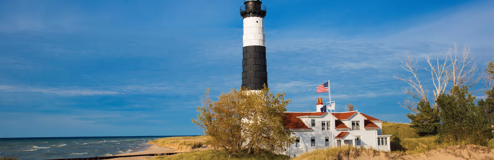 A black and white striped lighthouse next to a white building with a red roof, surrounded by trees and grassy dunes, with an American flag flying on a flagpole under a blue sky.