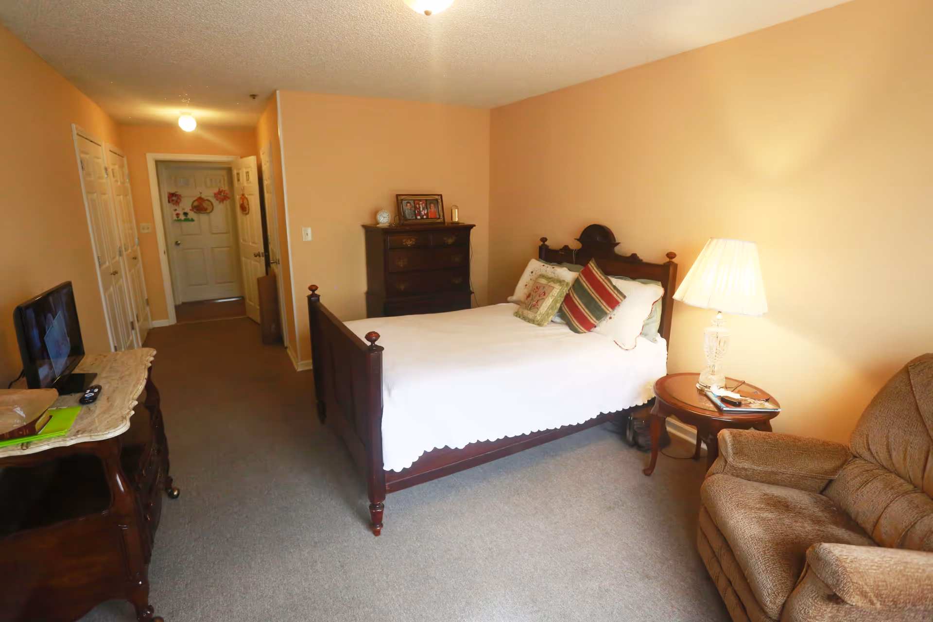 A furnished senior living facility bedroom with bed, dresser, sofa, and hallway.