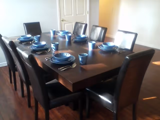 A dining room with a dark wooden rectangular table set for eight people. Each place setting includes a blue bowl, plate, cup, and silverware. The chairs around the table are black with cushioned backs and seats. The room has wooden flooring and light-colored walls with a closed white door in the background.