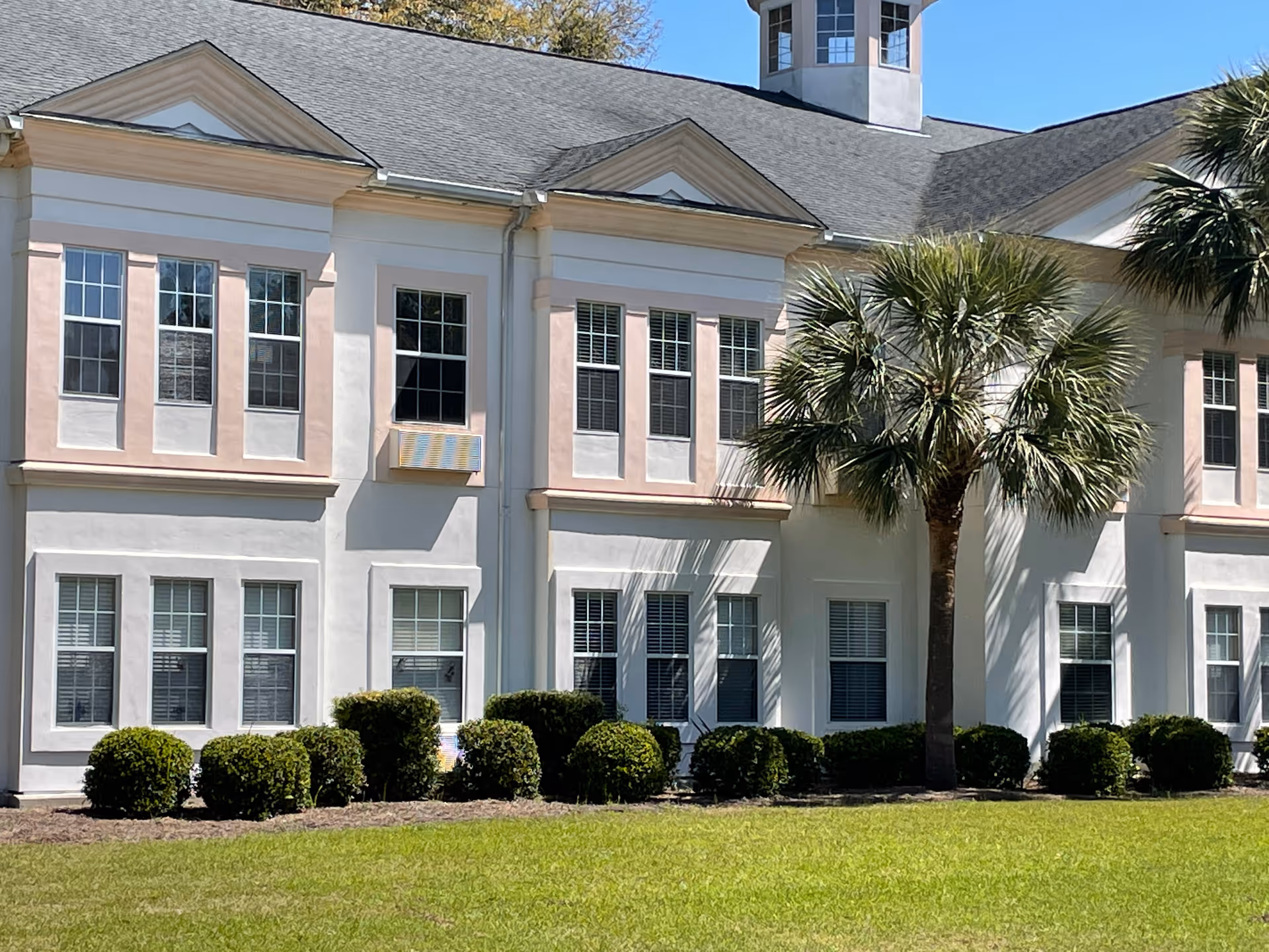 White two-story senior living building with pink trim, palm trees, and trimmed hedges.