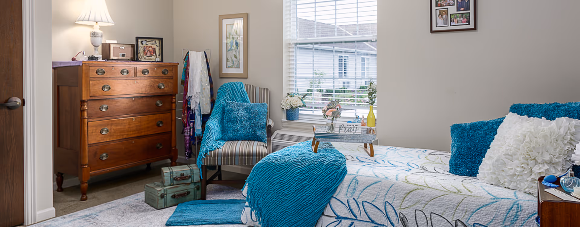 Sunlit bedroom with a bed dressed in blue-and-white linens, a wooden dresser, and a striped armchair by the window with decorative accents.