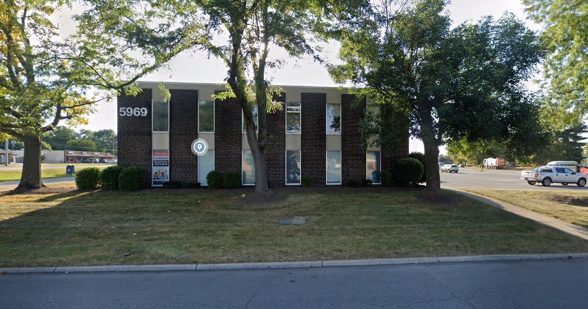 Front facade of a brick senior living facility with trees and parked cars.