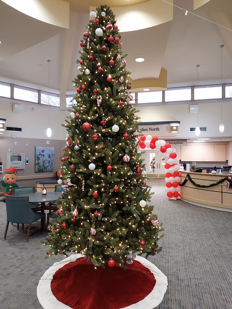 Tall Christmas tree with red and white ornaments in a facility lobby near a reception desk.