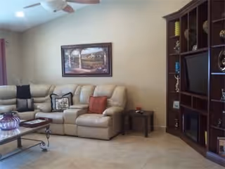 A living room with a beige sectional sofa adorned with several cushions, a glass coffee table with decorative items, a dark wooden entertainment center with a TV and shelves holding various objects, and a framed landscape painting on the wall above the sofa.