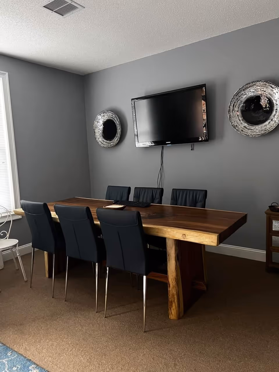A gray-walled dining room with a wooden table, black chairs, and a wall-mounted TV.