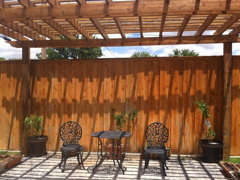 Outdoor patio with two ornate chairs, a metal table, potted plants, and a wooden fence.