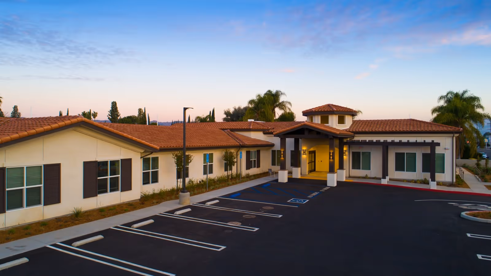 Front exterior of a single-story assisted living facility with tile roofs, an entrance canopy, empty parking spaces, and palm trees at dusk.