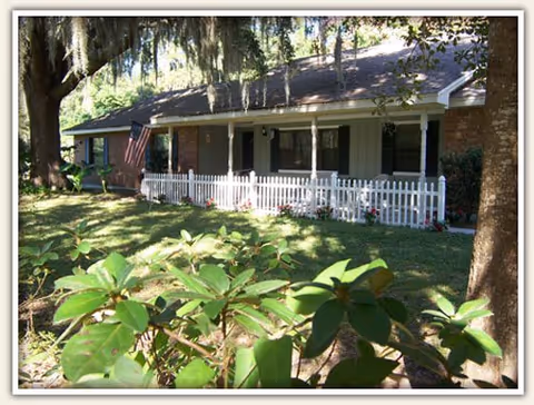 A single-story house with a white picket fence, front porch, and lush greenery in the foreground.