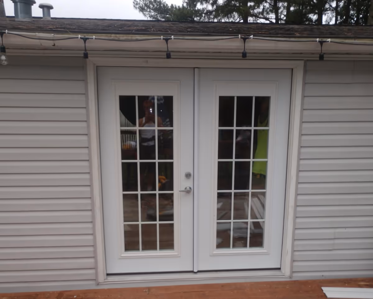 Exterior view of two white-framed French doors with grid panes on gray siding, string lights overhead.
