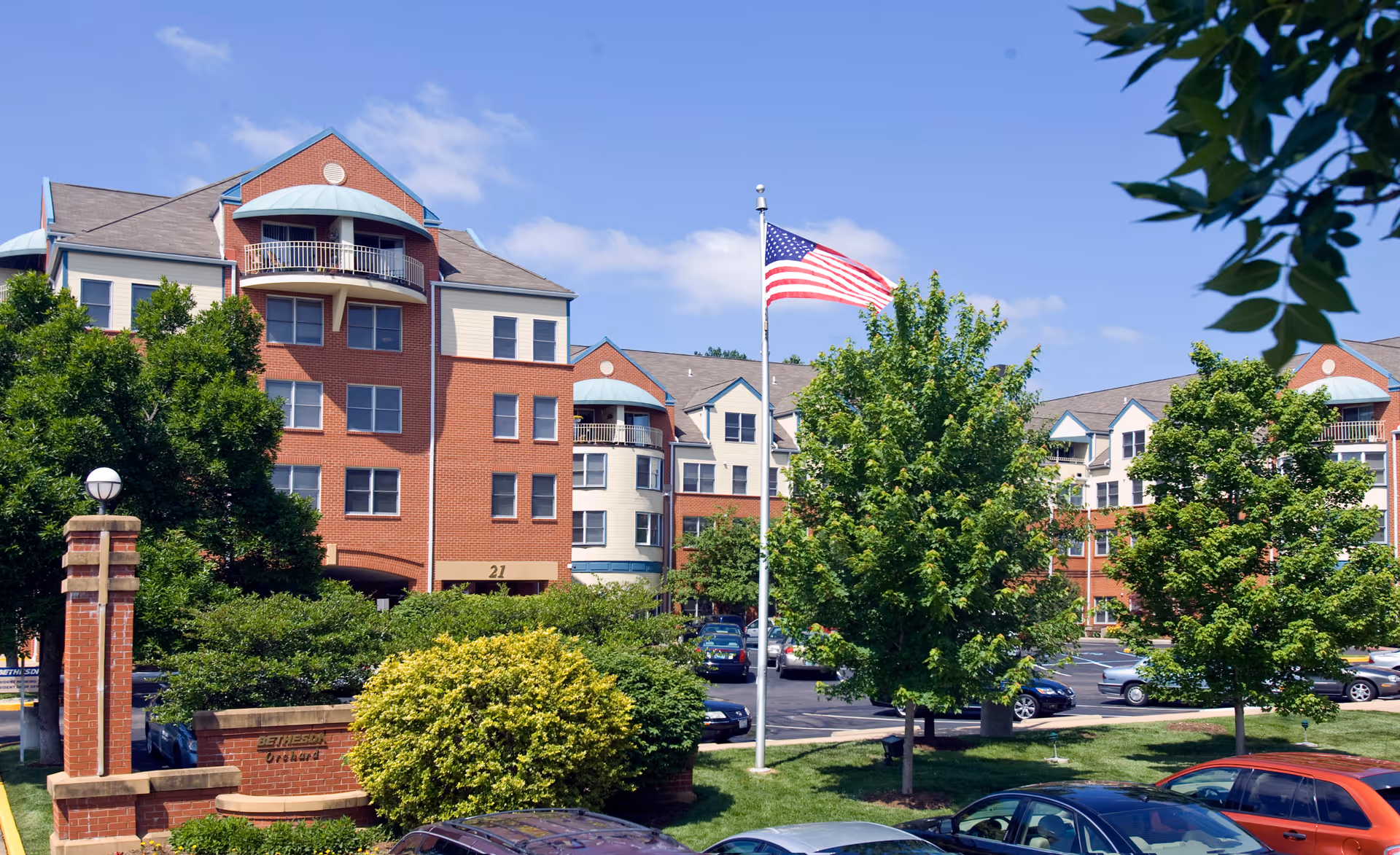 Front exterior of a multi-story brick senior living building with trees, an American flag, and parked cars.
