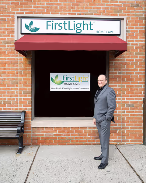 A man in a suit stands outside a FirstLight Home Care building under a red awning.