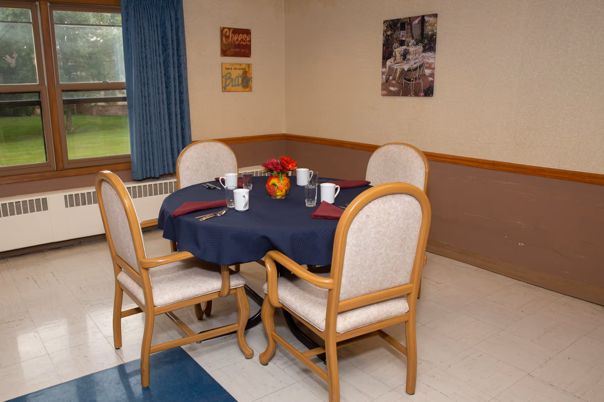 A small dining area with a round table covered by a dark blue tablecloth, set with four chairs. The table has four white mugs, four glasses, silverware, red cloth napkins, and a small floral centerpiece. The room has beige walls with a brown lower trim, a window with blue curtains, and two framed pictures on the wall.